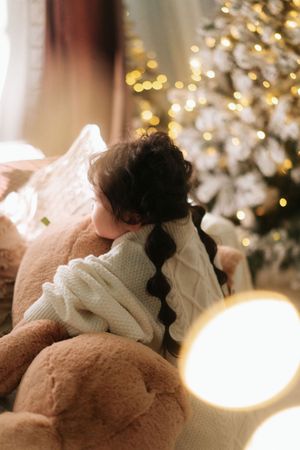 Child in white knit sweater hugging a teddy bear near a glowing Christmas tree with warm bokeh lights.