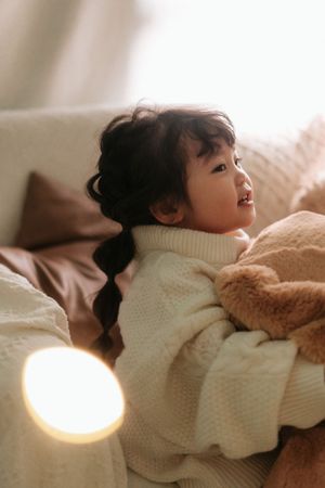 A young Asian girl in a white knit sweater cuddles a teddy bear on a cozy sofa, lit by warm lamplight.