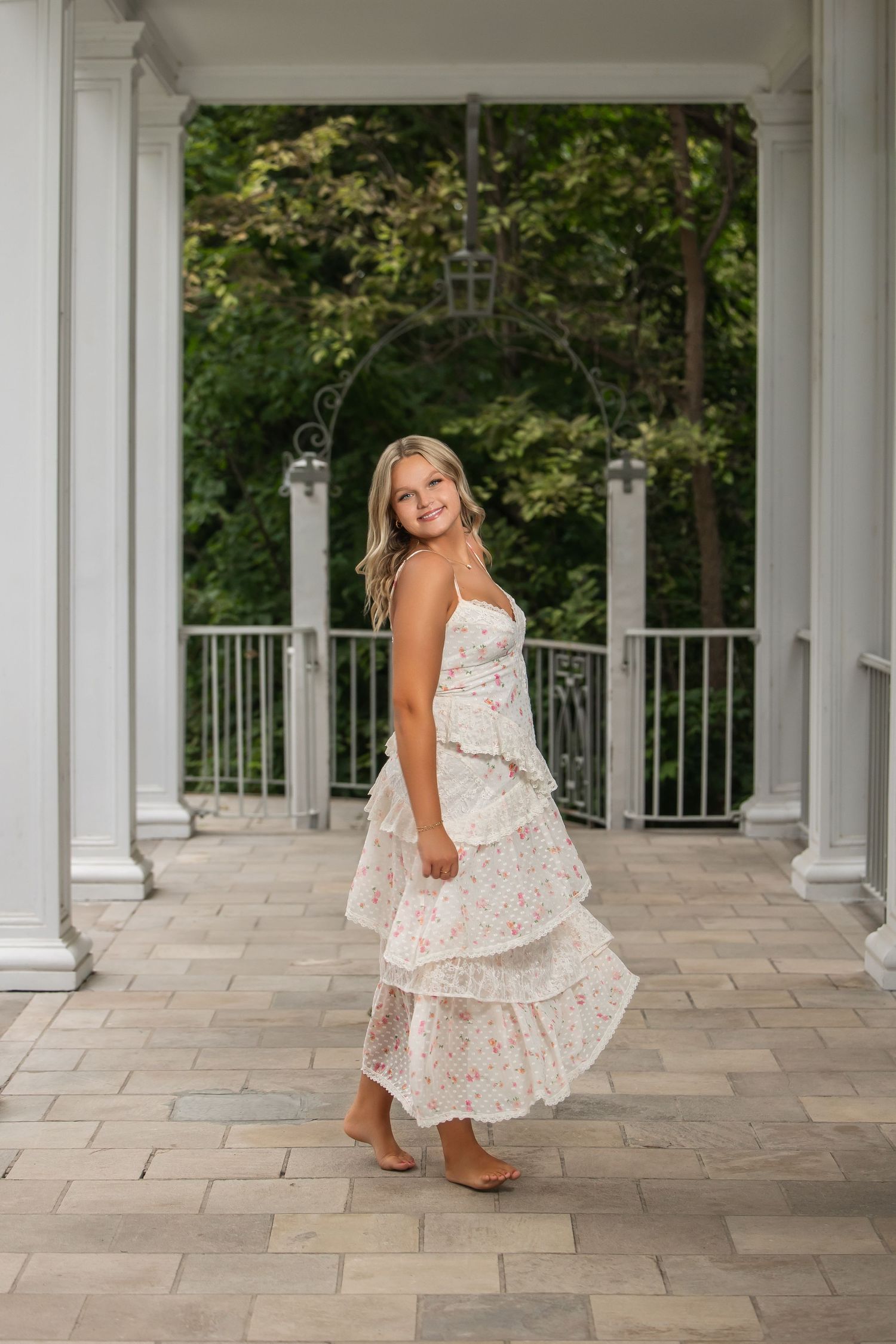 Young woman in floral tiered maxi dress posing barefoot on stone patio with white columns and lush greenery.
