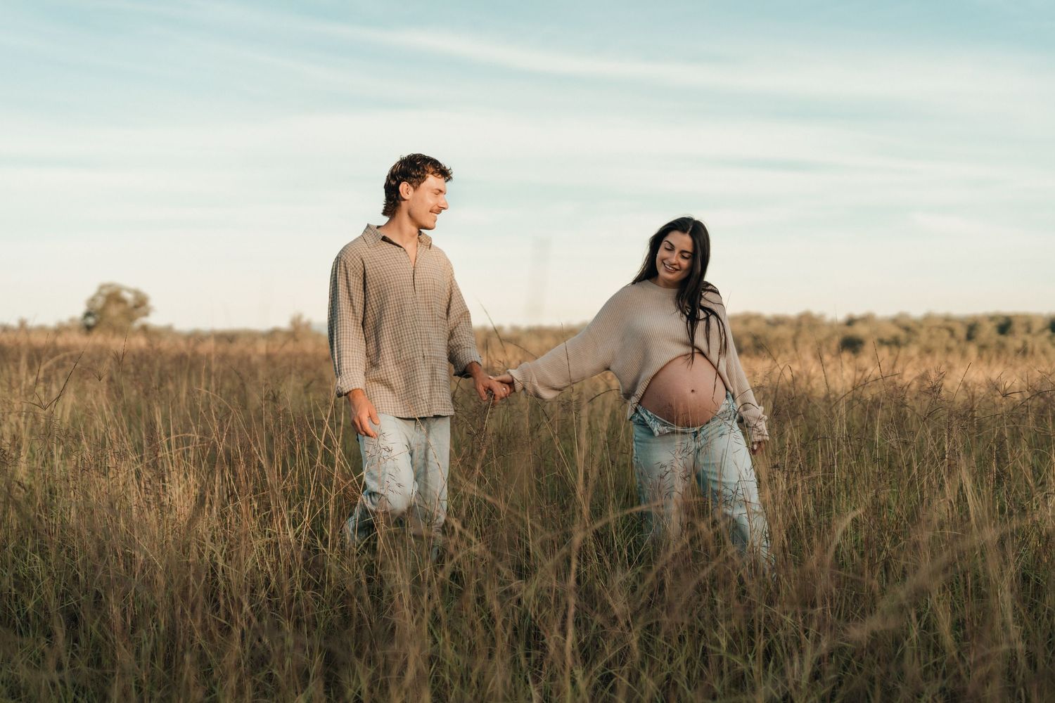 Pregnant woman in open jacket raises arms against blue sky, showing baby bump outdoors in natural light.