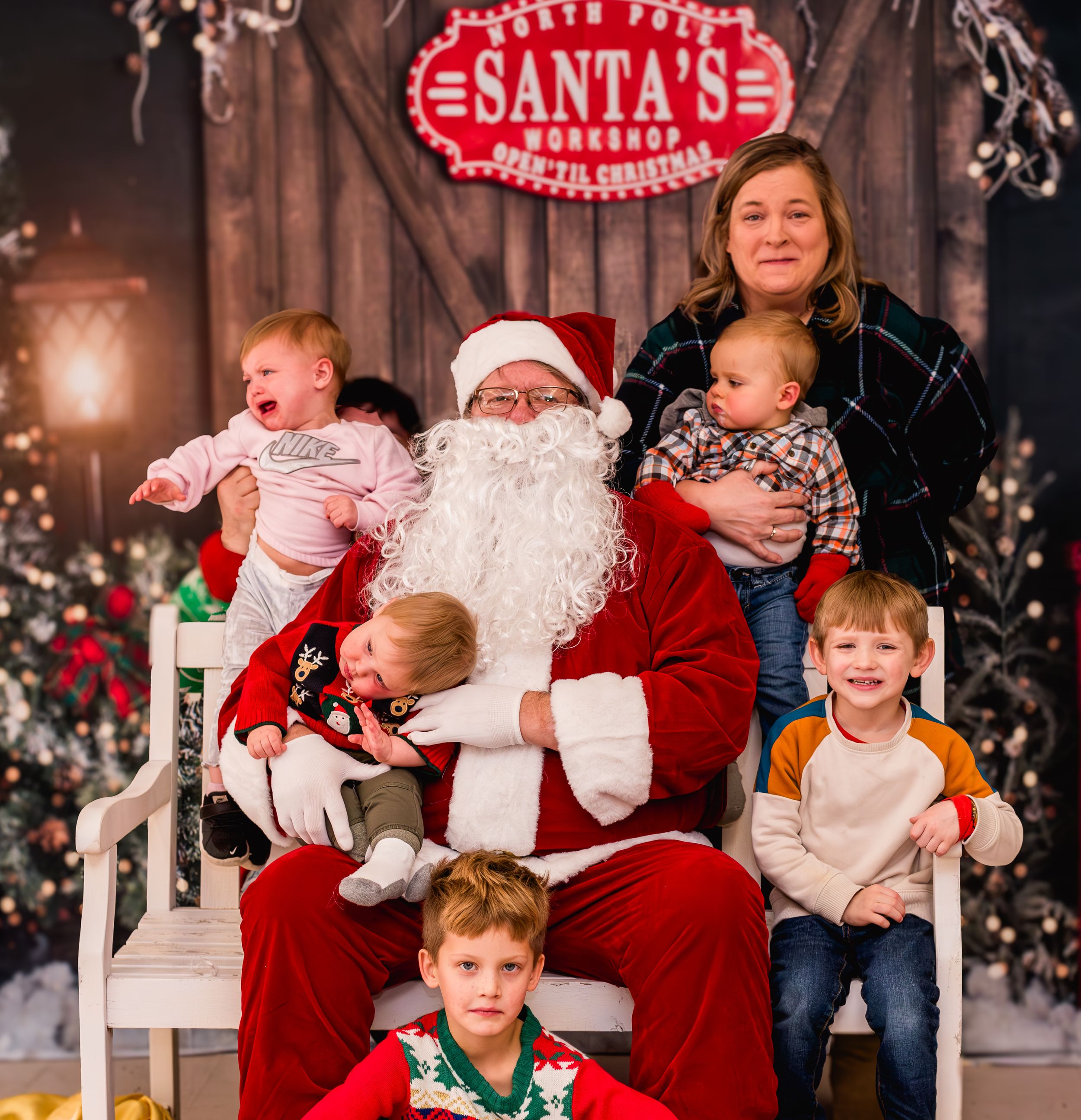 Two children sit with Santa Claus in his workshop, surrounded by festive Christmas decorations and trees.