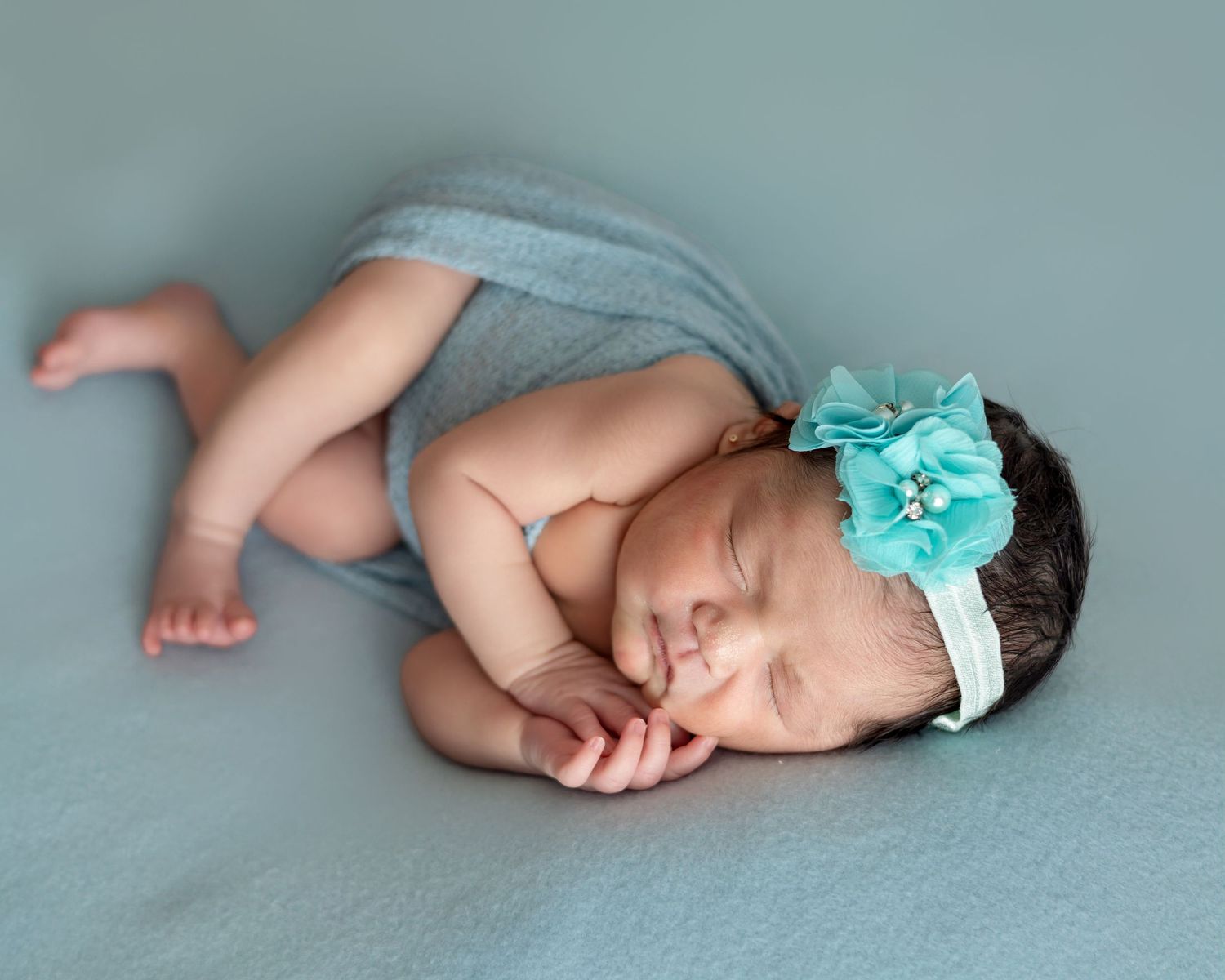Sleeping newborn baby girl wearing a teal flower headband, curled up on a soft blue blanket.