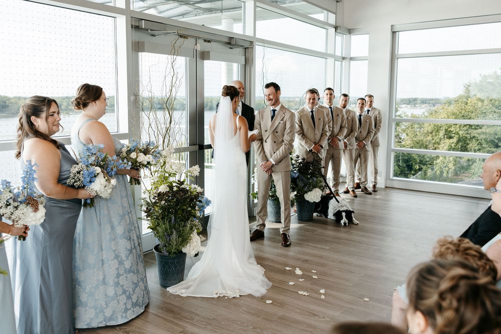 Wedding ceremony in a bright venue with bridesmaids in blue, groomsmen in tan suits, and couple exchanging vows by large windows.