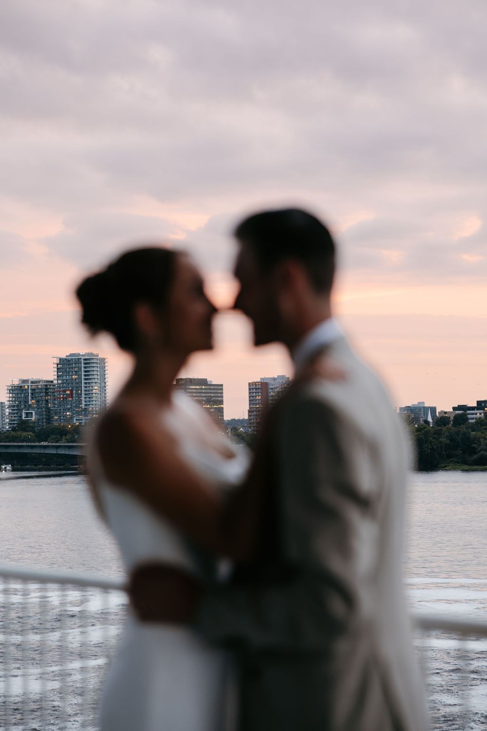 Bride and groom share a romantic moment by the waterfront at sunset with a city skyline in the background.