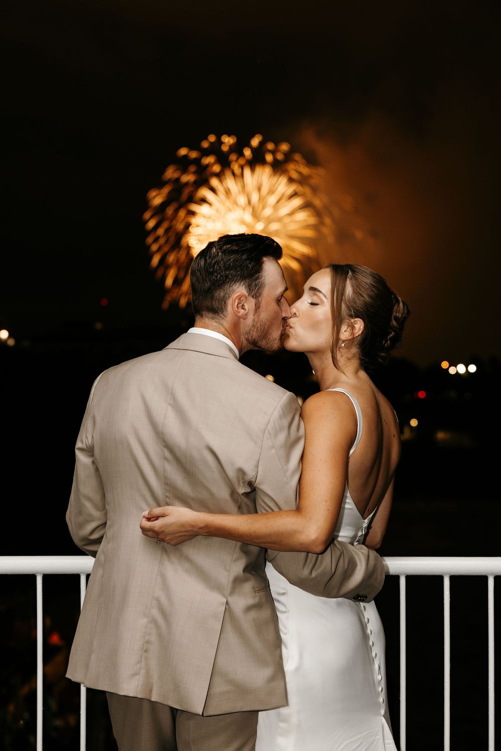 Bride and groom share a romantic kiss on a balcony with golden fireworks lighting the night sky behind them.