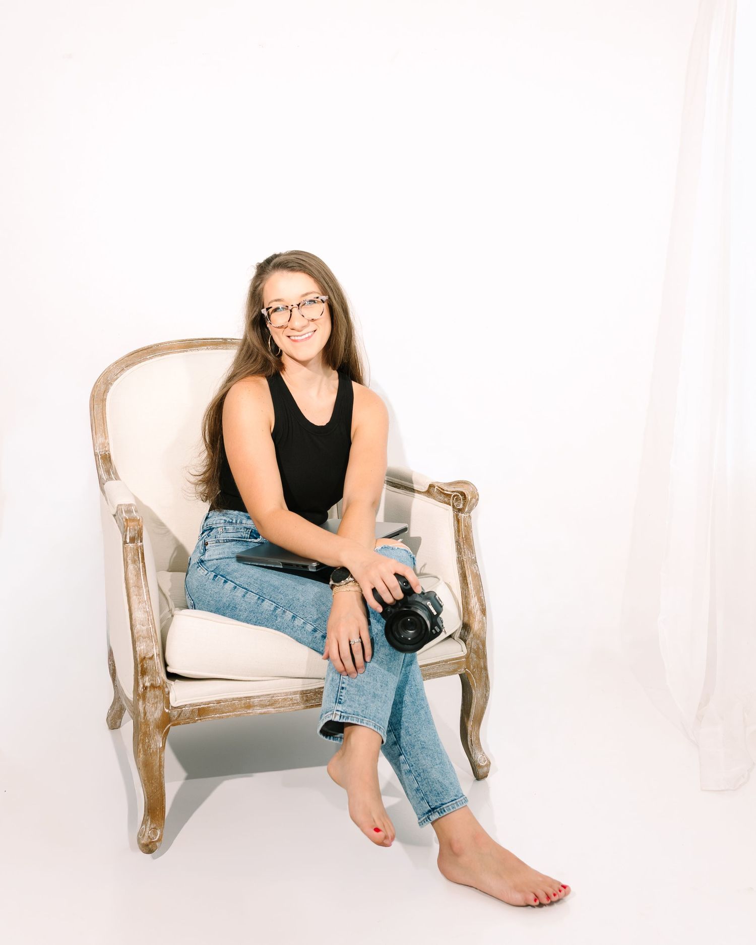 Woman photographer sitting barefoot in vintage chair holding camera, wearing jeans and black top, smiling in bright studio.