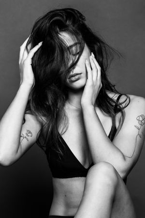 Black and white portrait of a tattooed woman in a bralette with hair covering her face, hands on head.