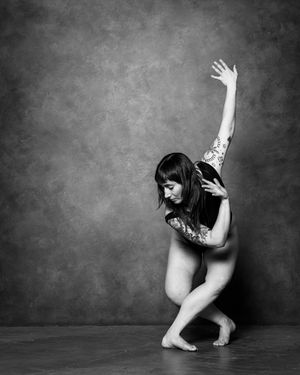 Black and white photo of a nude woman with tattoos striking a dramatic dance pose against a textured backdrop.