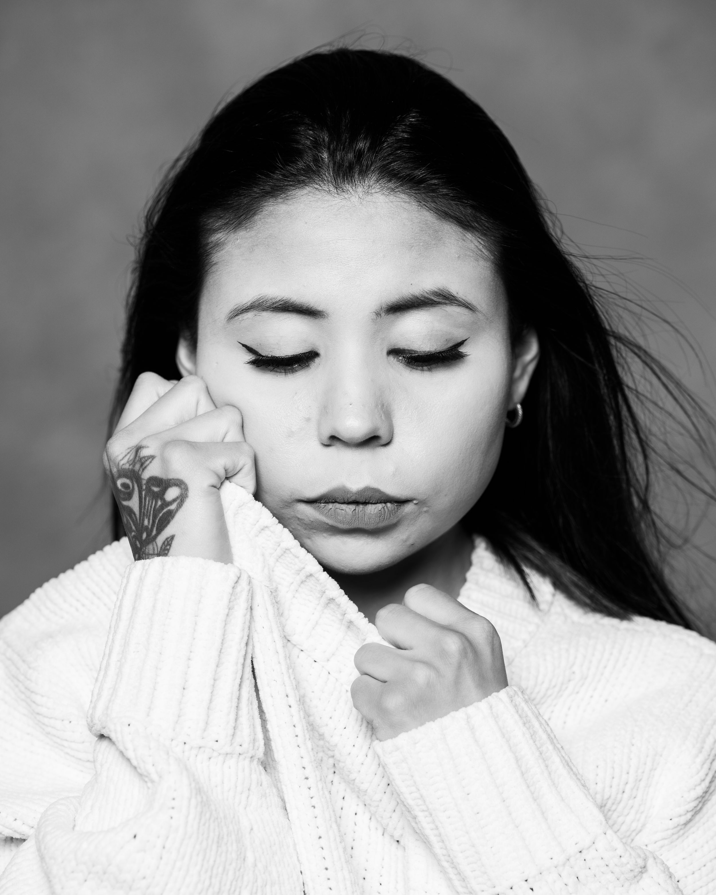 Black and white close-up of a woman with closed eyes, resting her tattooed hand against her cheek.