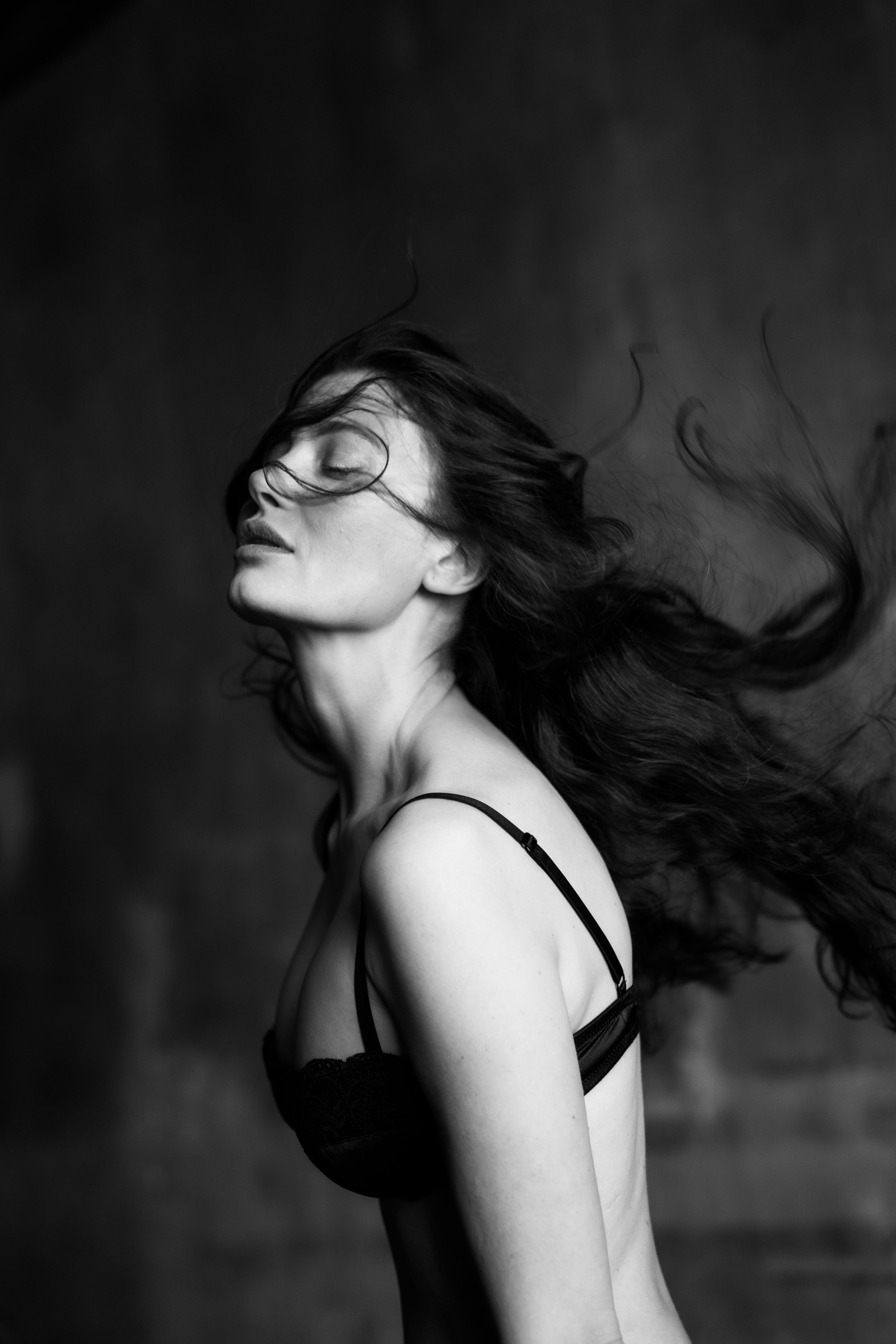Black and white portrait of a woman with long dark hair blowing across her face in a dark setting.