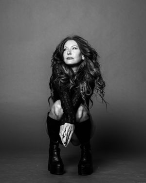 Black and white photo of a woman with long wavy hair crouching in boots, looking upward dramatically.
