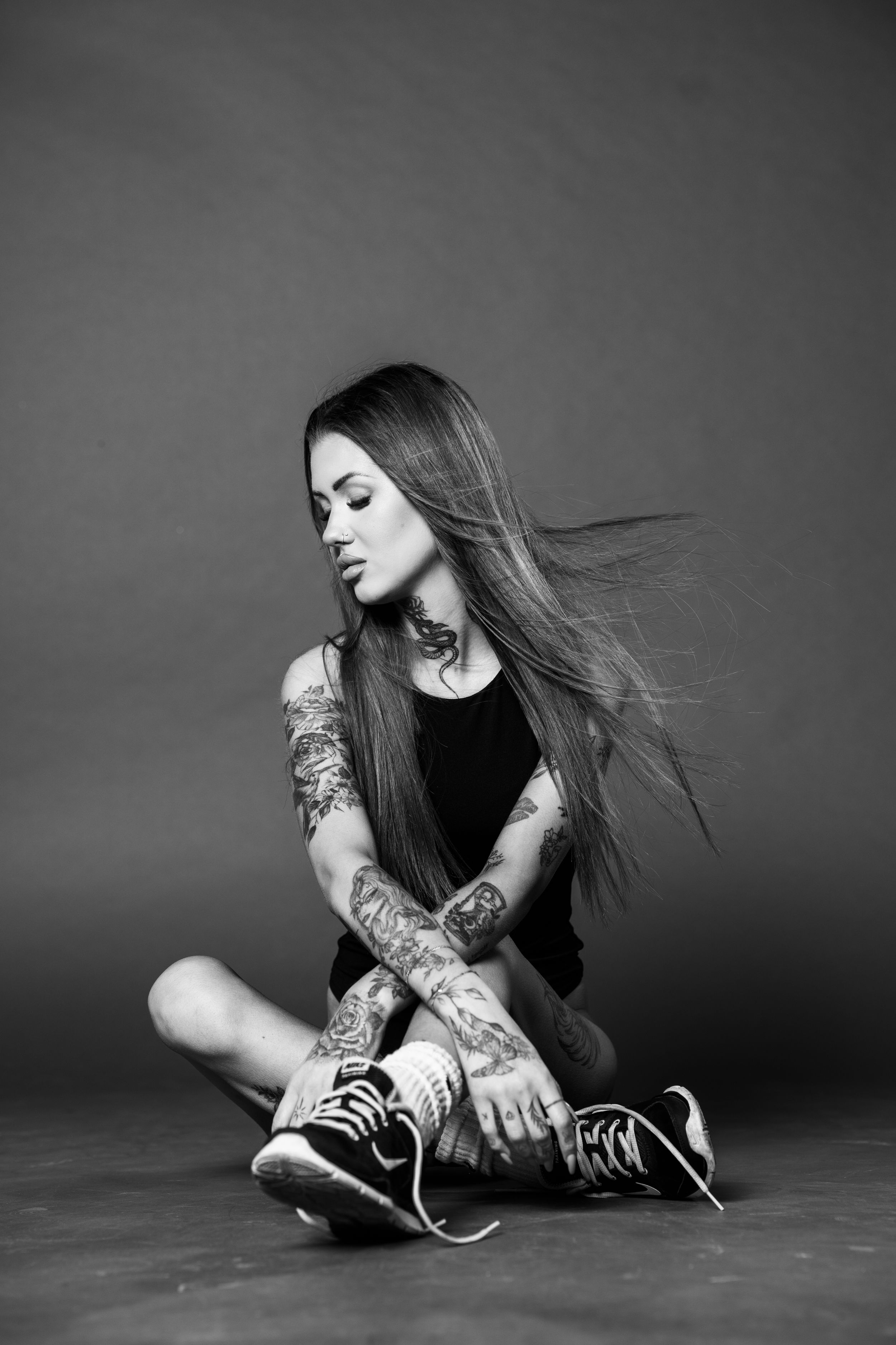 Black and white photo of a heavily tattooed woman sitting cross-legged with her hair blowing sideways.