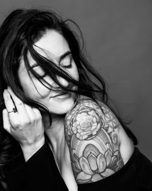Black and white portrait of a woman with flowing hair and floral shoulder tattoo, eyes closed, hand near face.