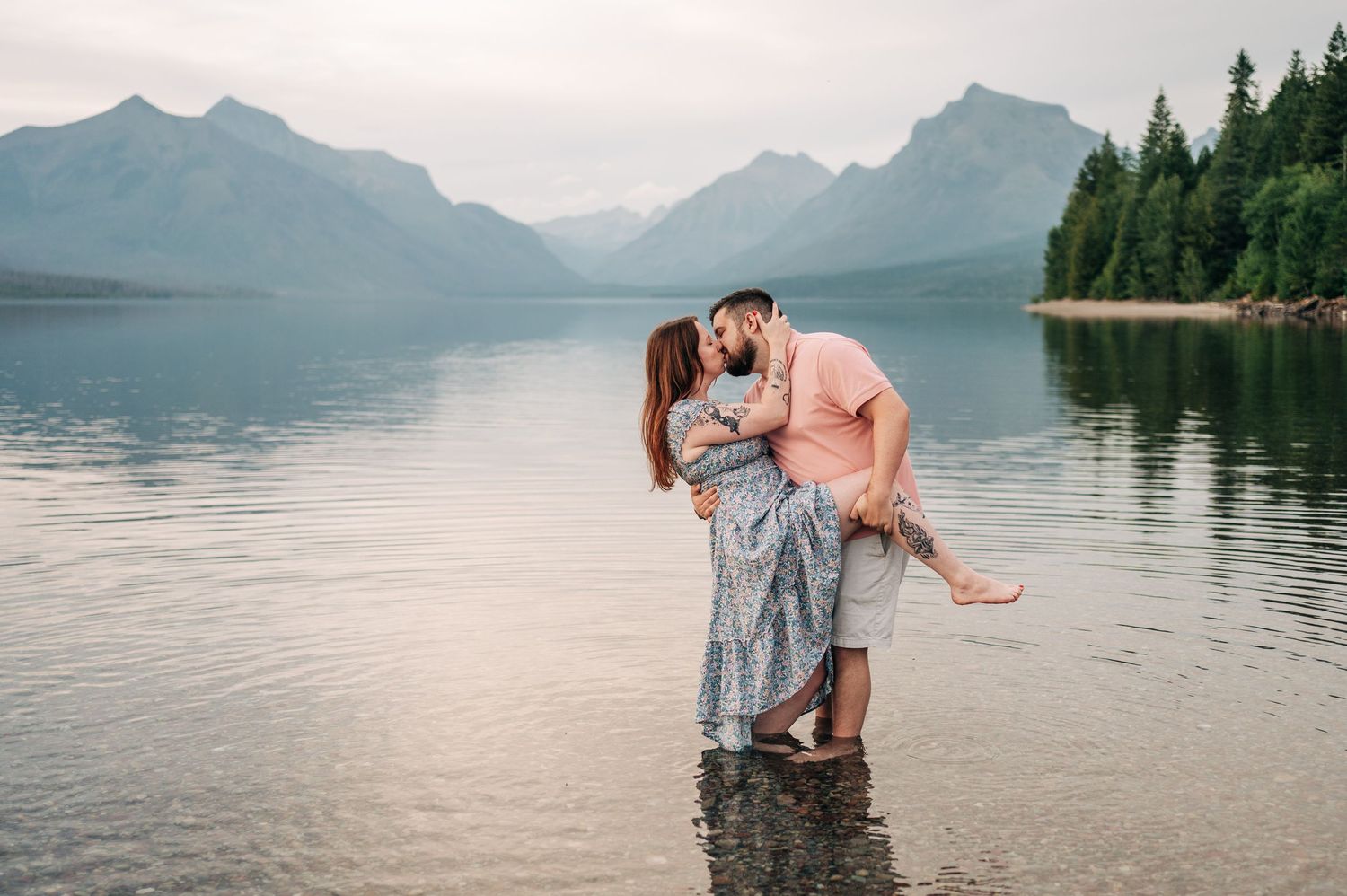 Couple sharing a romantic kiss while standing in a serene mountain lake with misty peaks and evergreen trees.