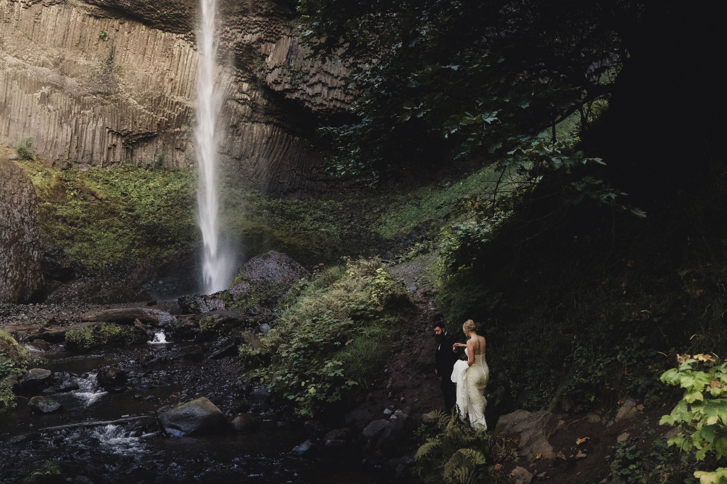Couple embracing near a stunning waterfall surrounded by lush green moss-covered rocks in a forest canyon.
