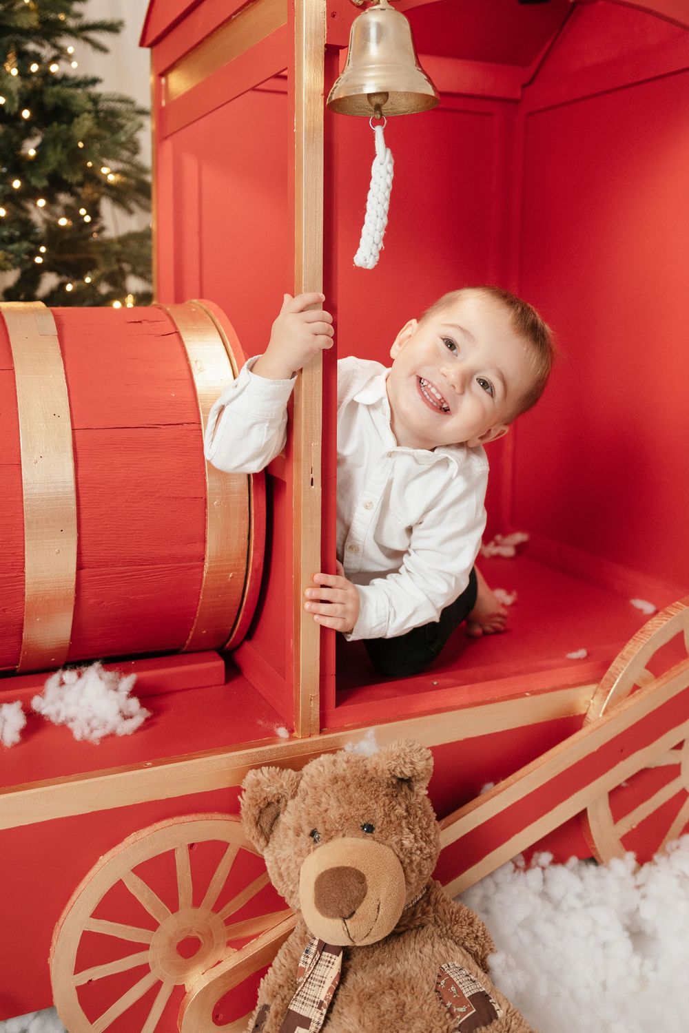 Smiling boy in white shirt plays inside a red toy train with a bell, teddy bear, and Christmas tree in background.