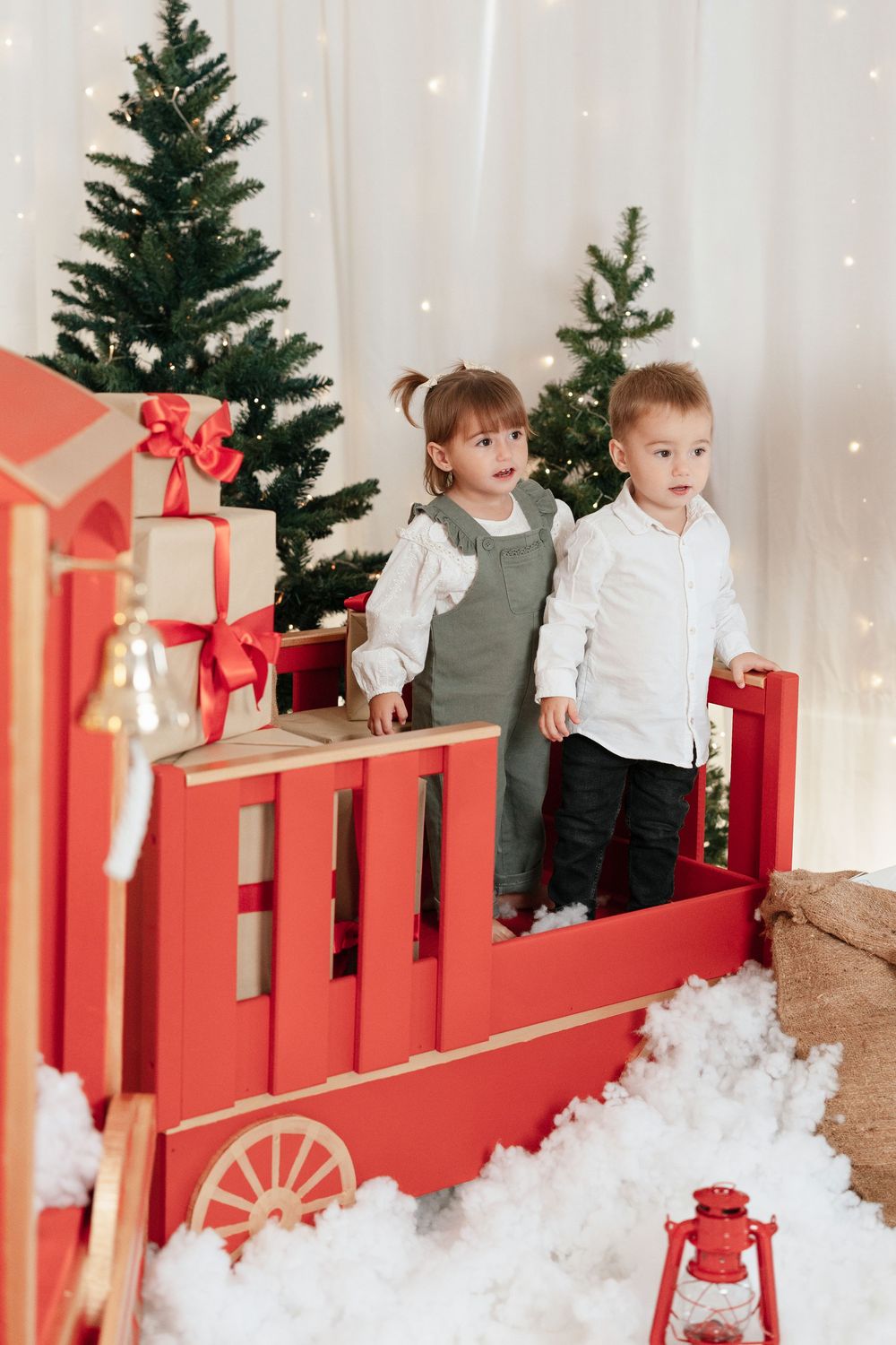 Two young children stand in a red toy train car surrounded by Christmas trees and gifts in a festive holiday setting.