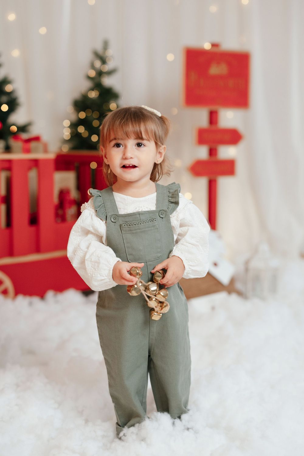 Toddler girl in sage green overalls holds gold bells in a festive Christmas photo studio with tree and red gifts.