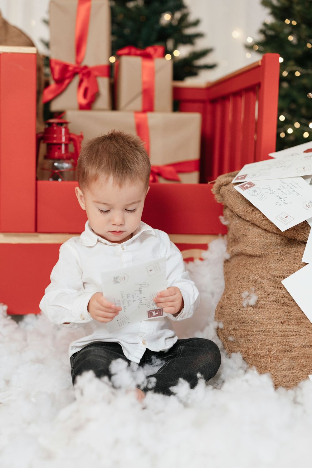 Young boy in white shirt reads letter sitting near Christmas gifts, burlap sack, and decorated tree with bokeh lights.