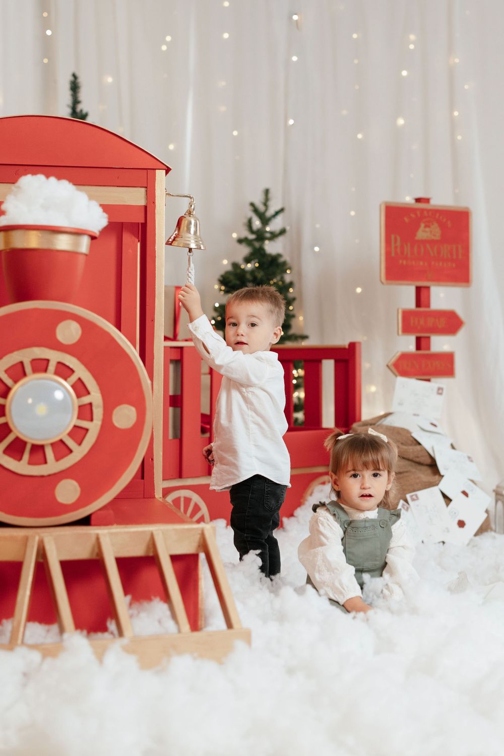 Two young children pose beside a large red toy train in a festive Christmas setting with snow and holiday decorations.