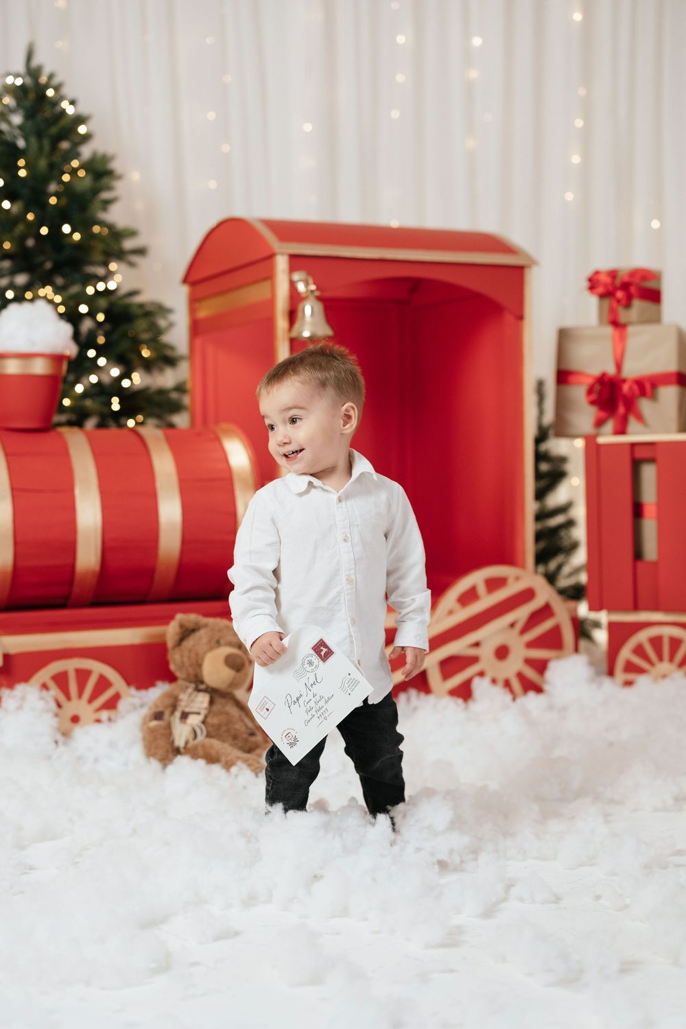 Smiling toddler boy in white shirt holding letter and teddy bear beside red toy train in Christmas holiday scene.