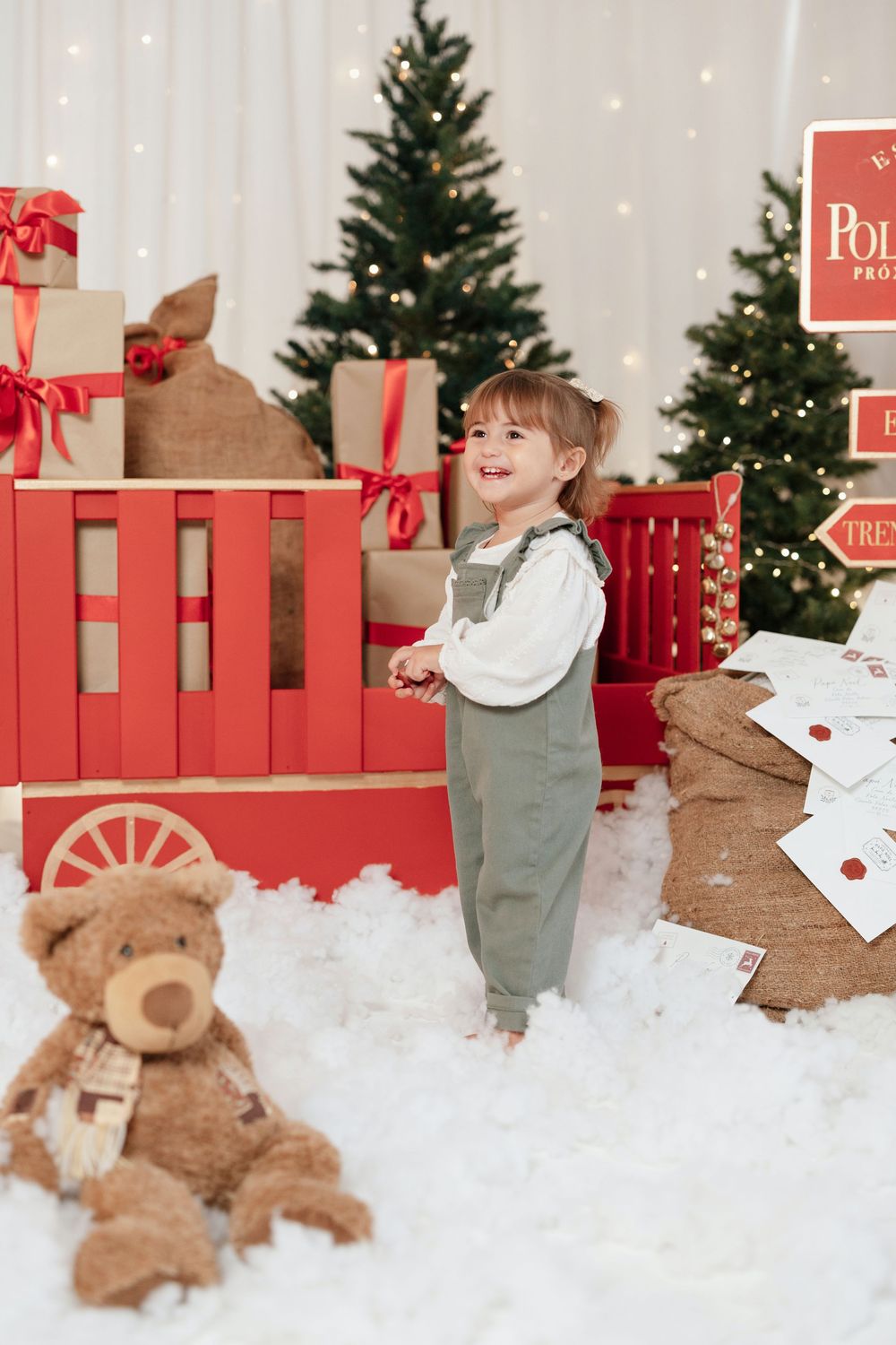 Smiling toddler in green overalls stands by a red Christmas wagon with gifts, teddy bear, and decorated trees in background.
