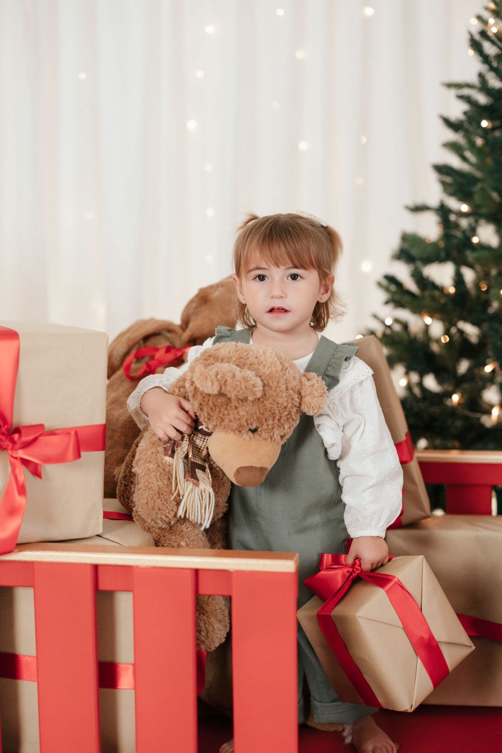 Toddler girl holding teddy bear surrounded by Christmas gifts near decorated tree with warm bokeh lights.
