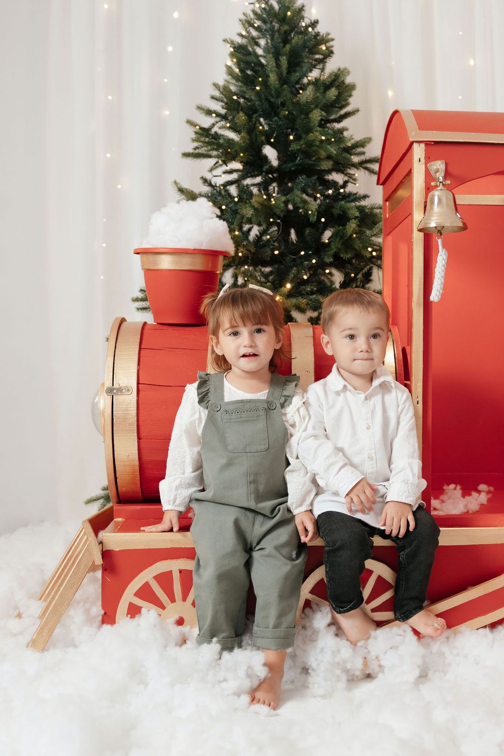 Two young children sit in front of a decorated Christmas tree beside a large red toy train in a festive holiday setting.