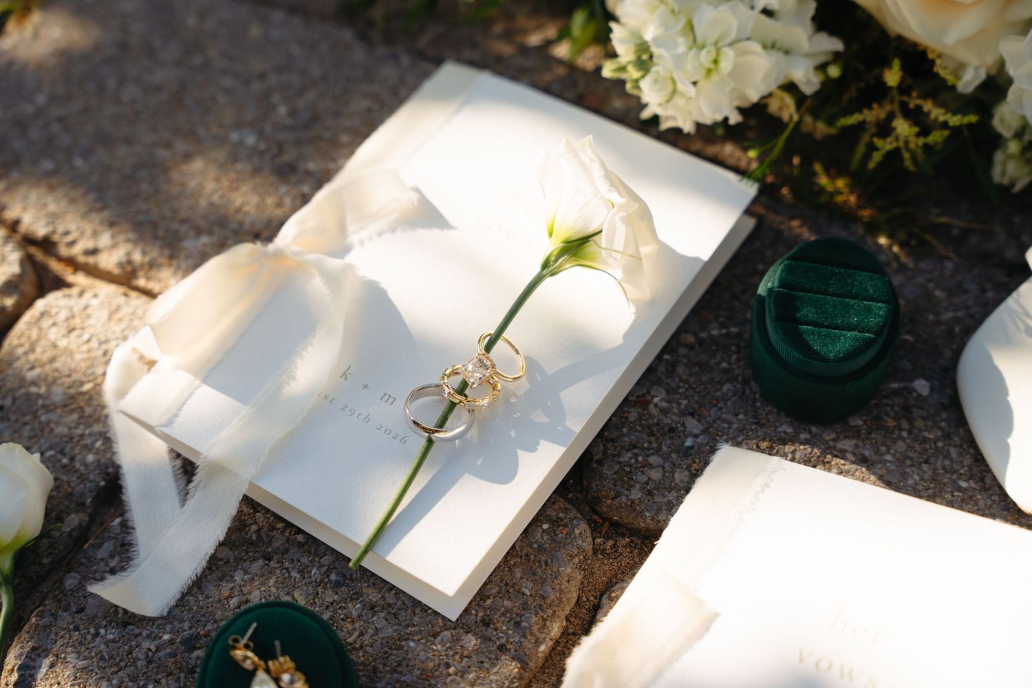Wedding rings resting on white ribbon and floral stationery with a green velvet ring box nearby.