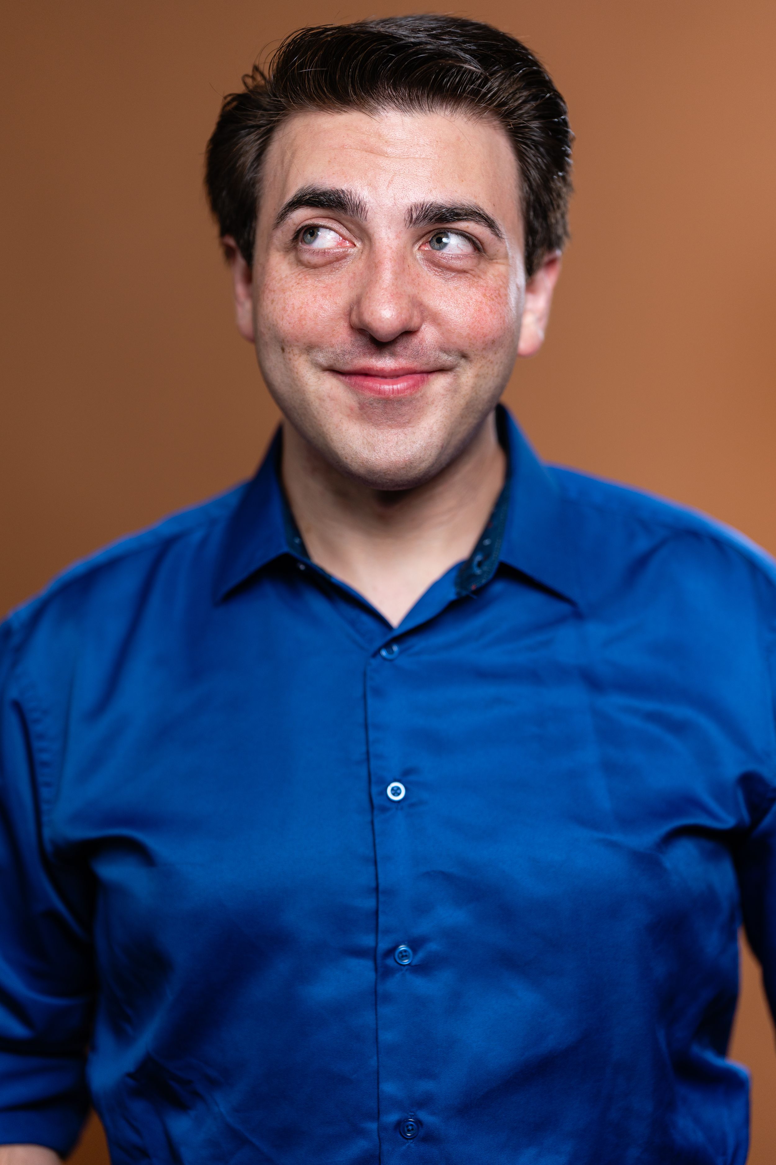 A young man in a blue dress shirt smirks and glances sideways against a brown studio background.