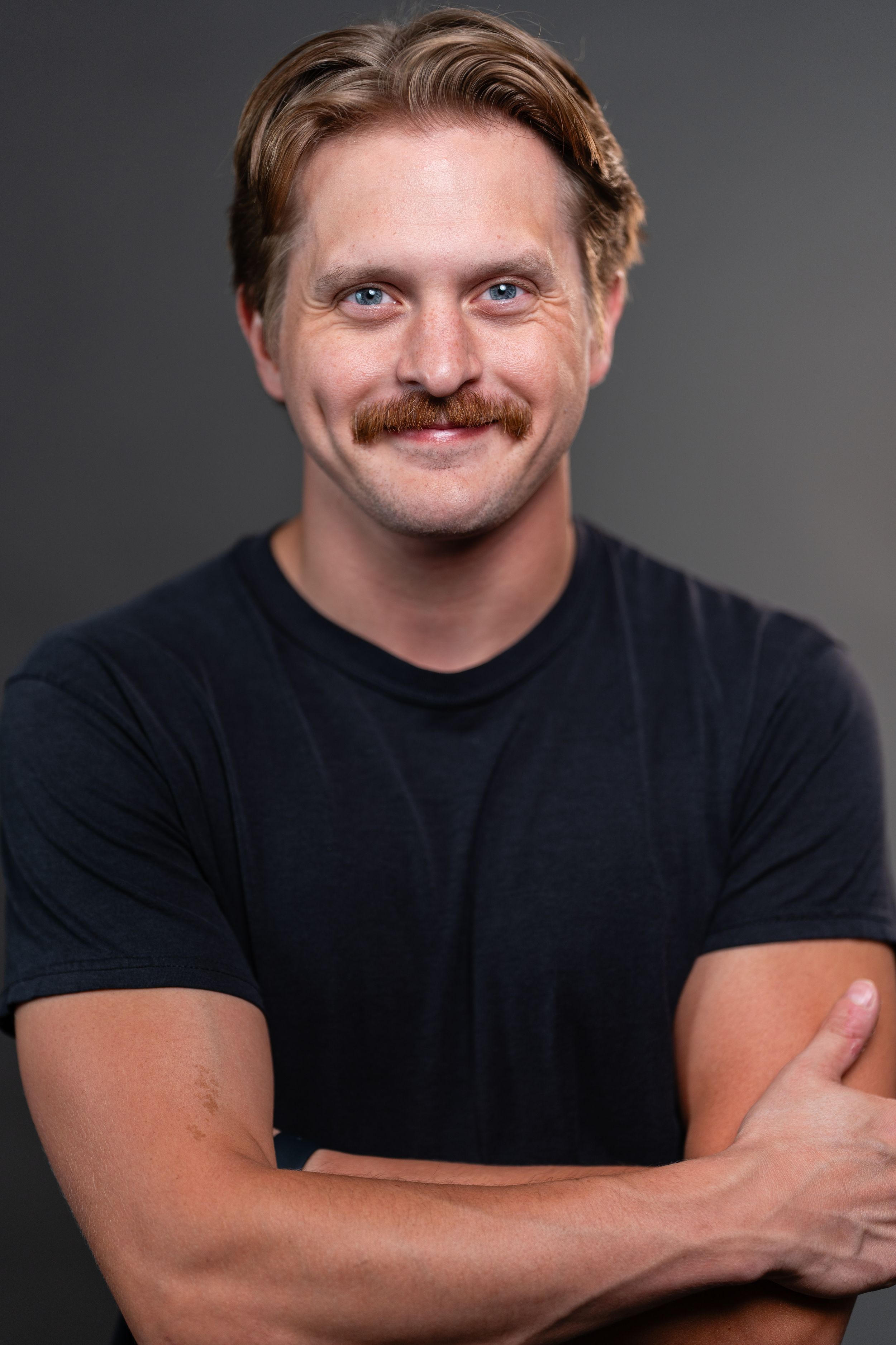A smiling man with a mustache and blue eyes, wearing a black t-shirt, arms crossed, against a gray background.