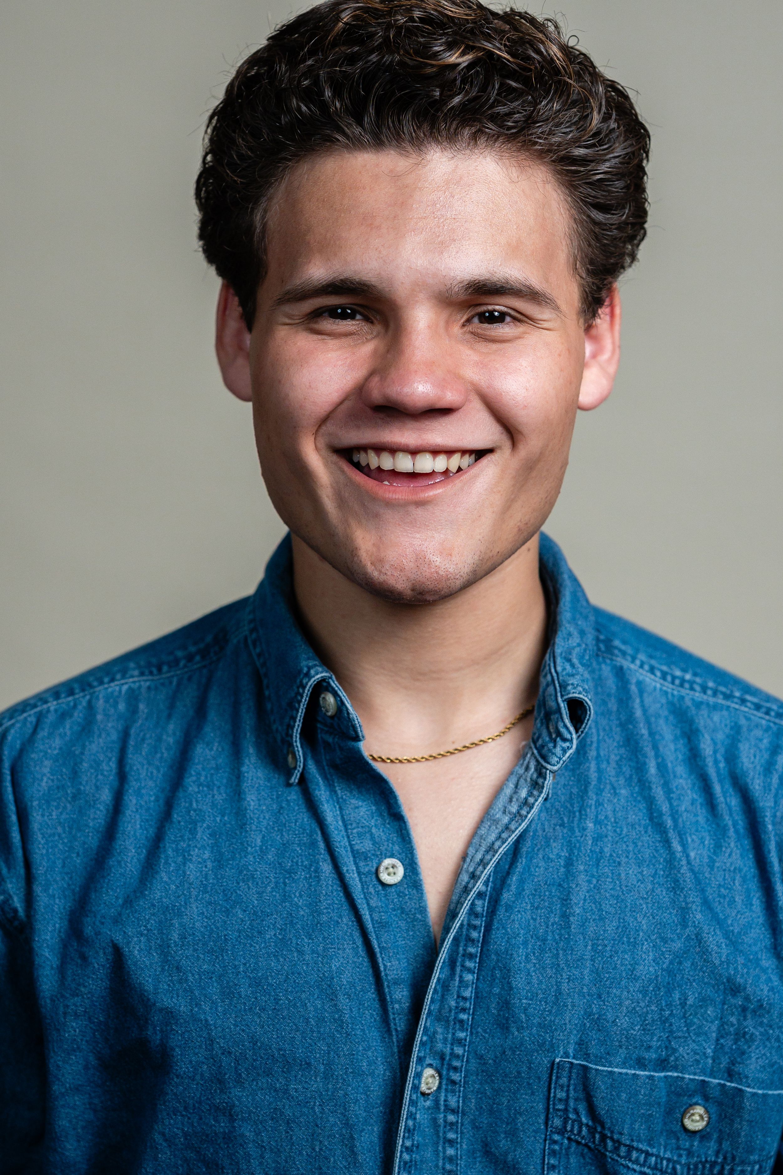 Smiling young man in a denim shirt and gold chain necklace posing for a portrait photo.