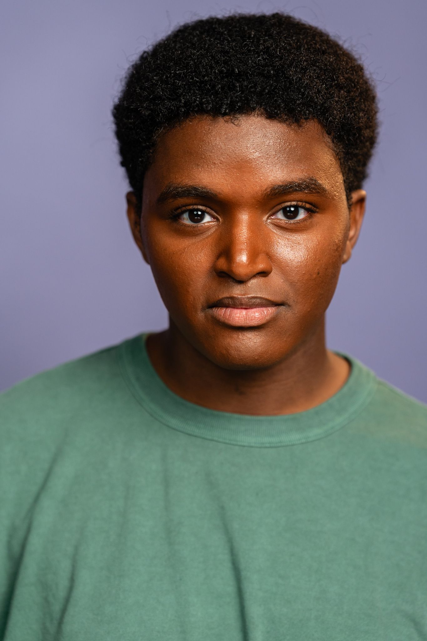Young Black man with natural hair wearing a green t-shirt poses for a portrait against a purple background.
