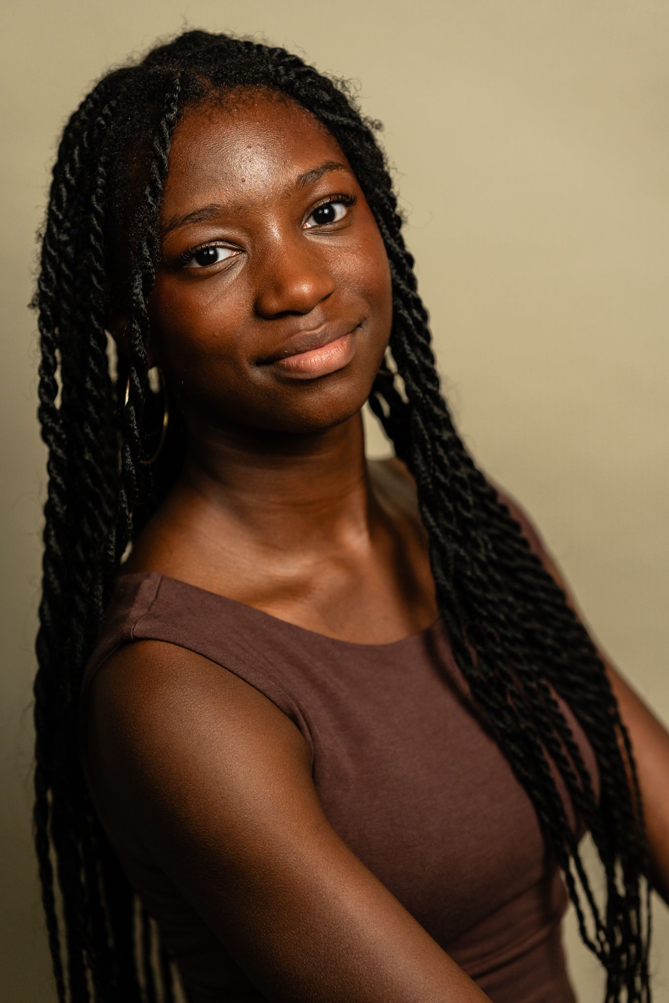Young woman with long braided hair wearing a brown sleeveless top, smiling softly against a neutral background.