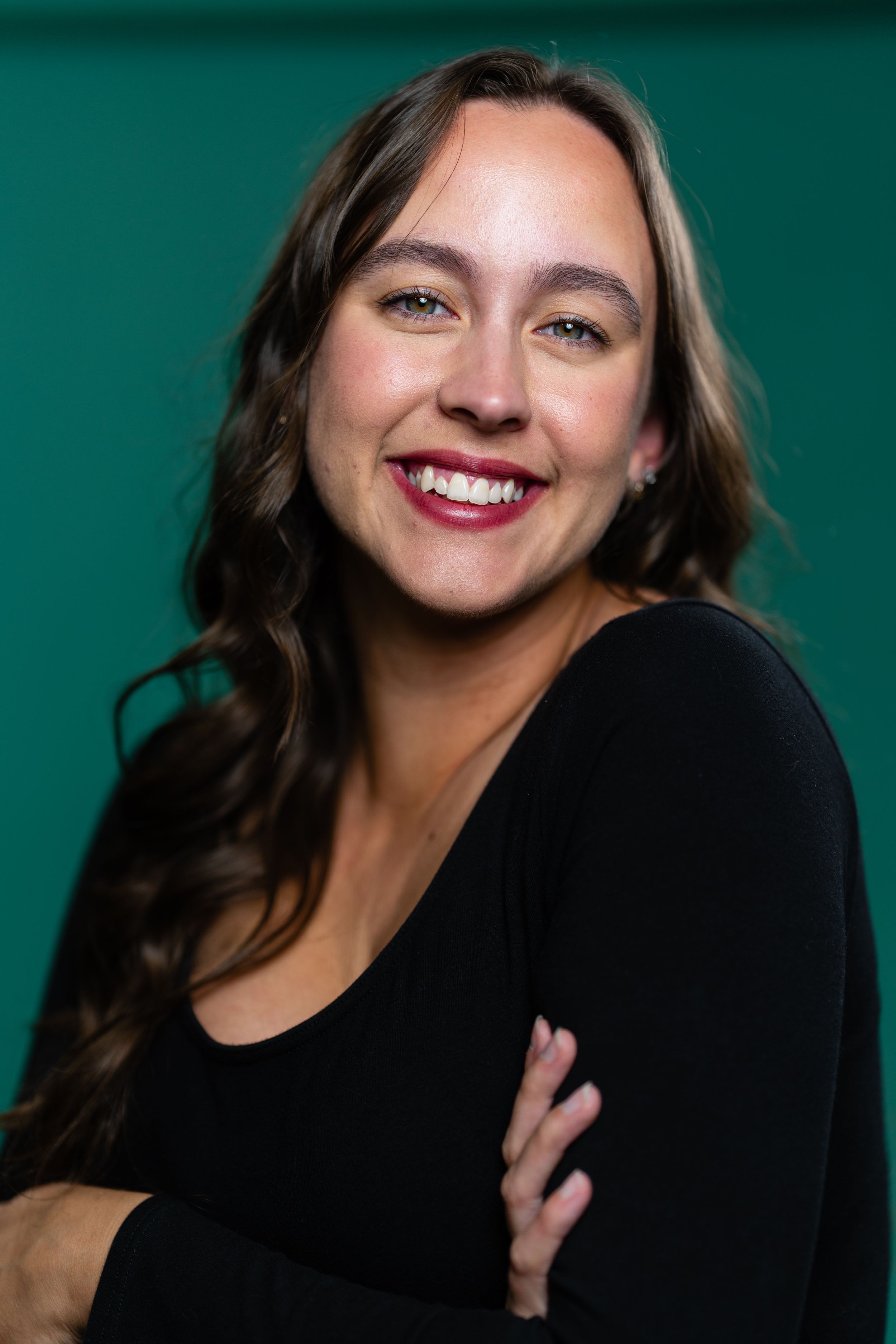 Smiling woman with long wavy brown hair in black top, arms crossed, posing against a teal green background.