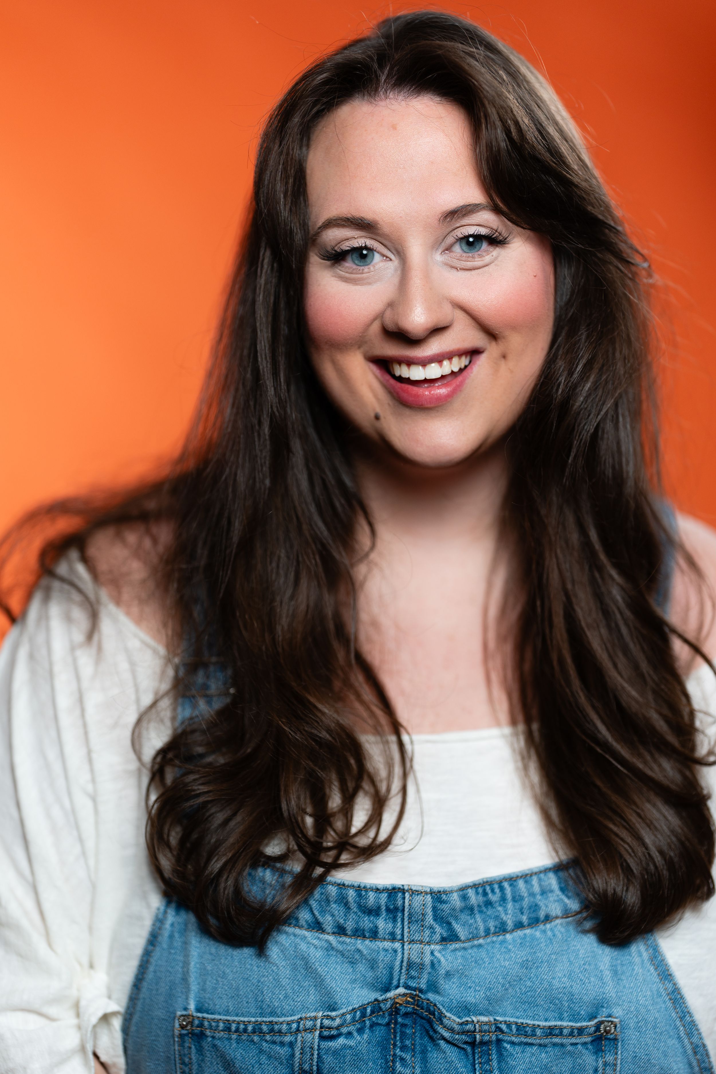 Smiling young woman with long brown hair wearing denim overalls and white shirt against orange background.