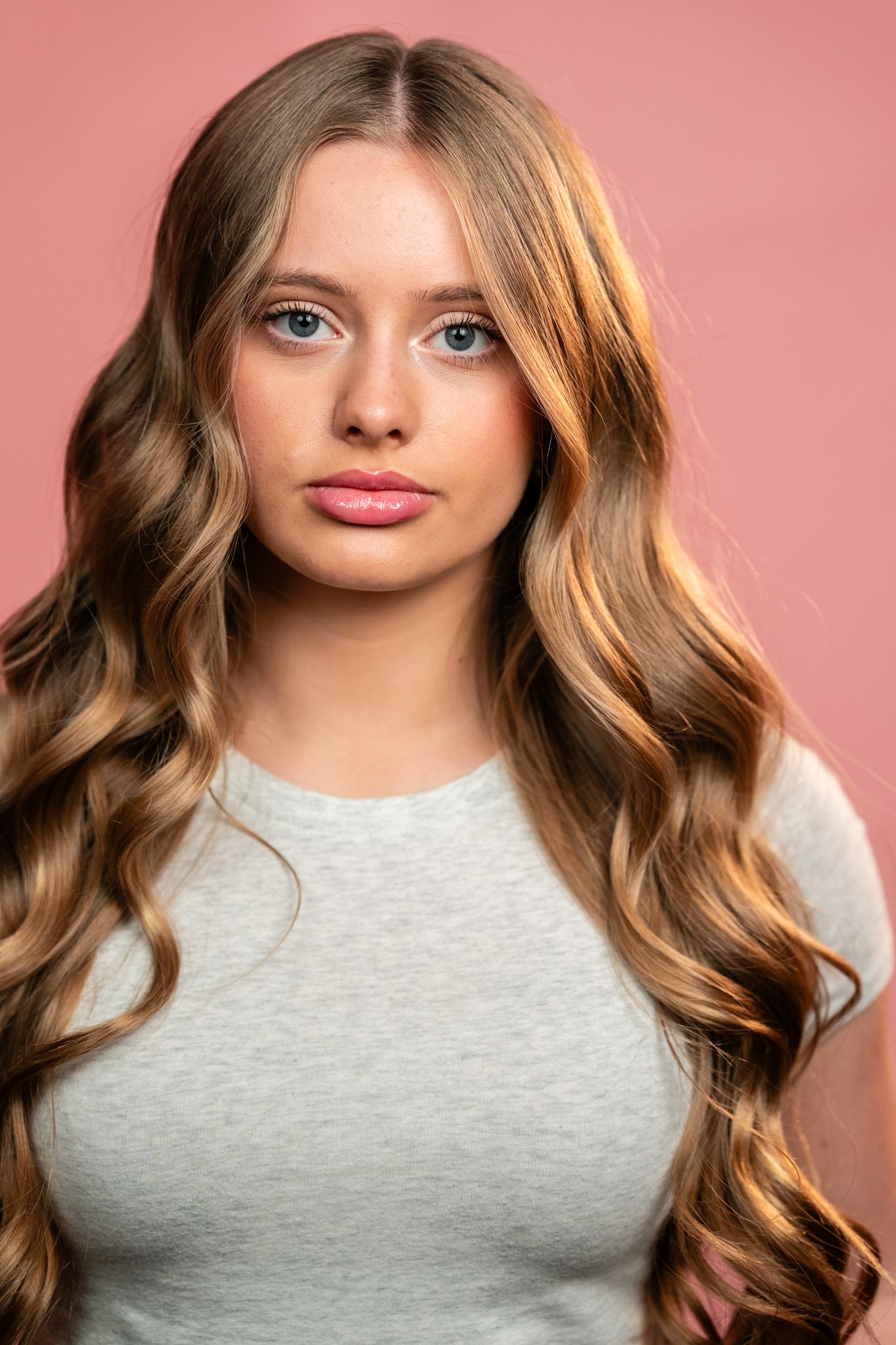 Young woman with long wavy blonde hair and blue eyes wearing a gray top against a pink background.
