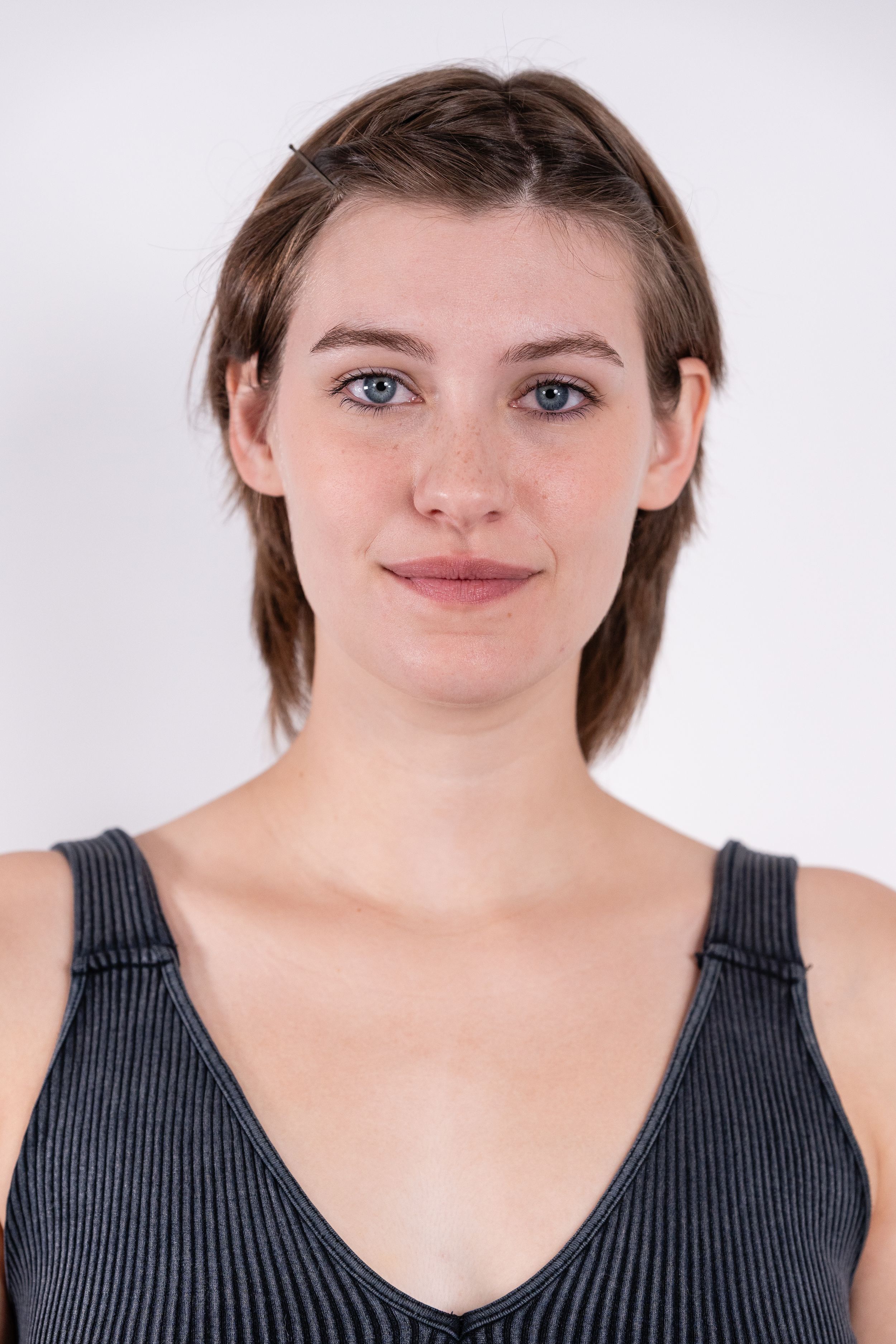 Young woman with short brown hair and blue eyes wearing a gray tank top, posing against a white background.