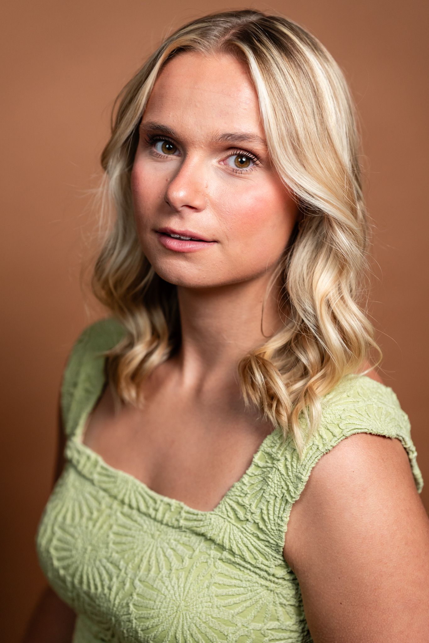 A blonde woman in a green lace top poses against a brown background in a professional portrait photo.