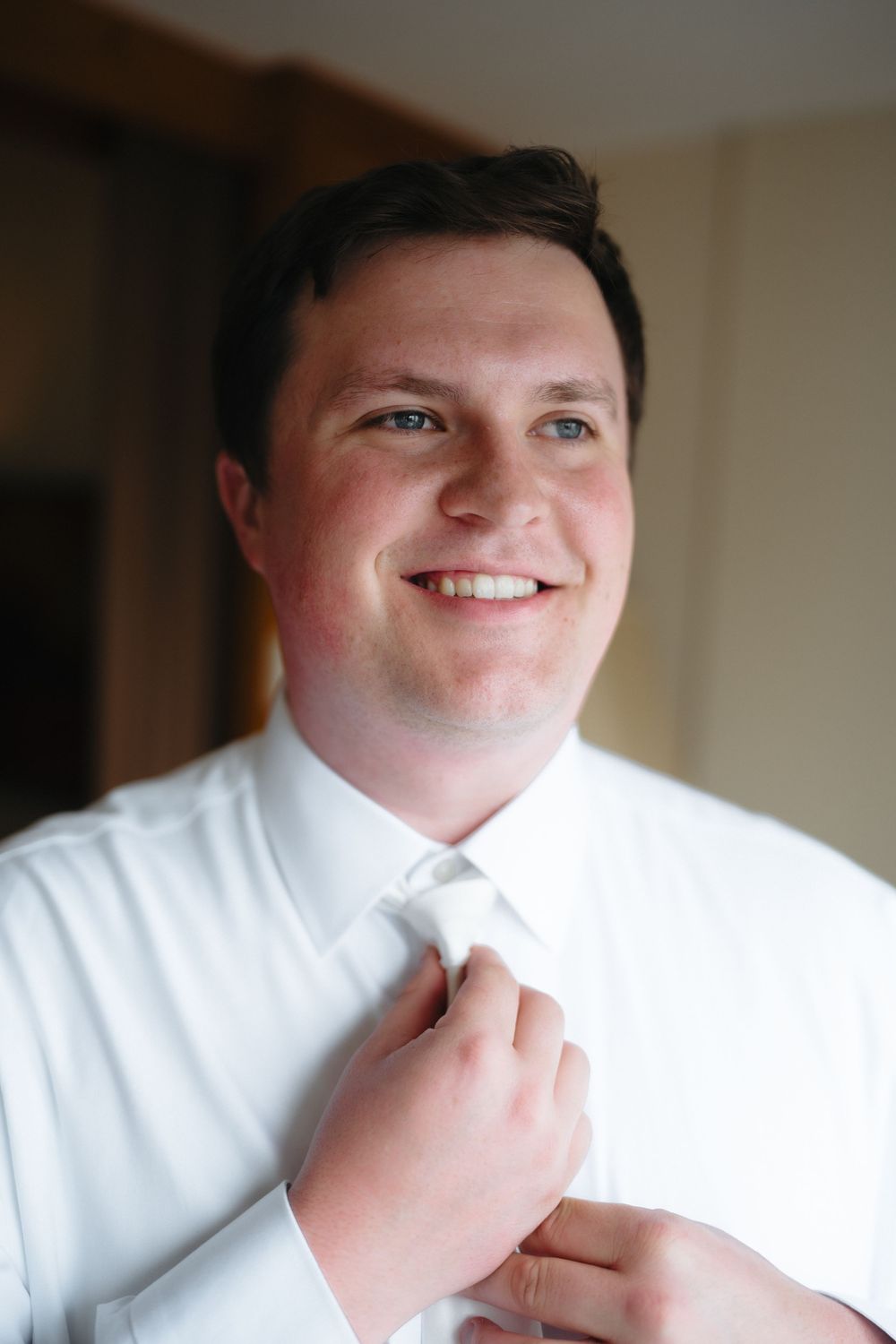 Smiling groom in white shirt adjusting his tie while getting ready for his wedding day.