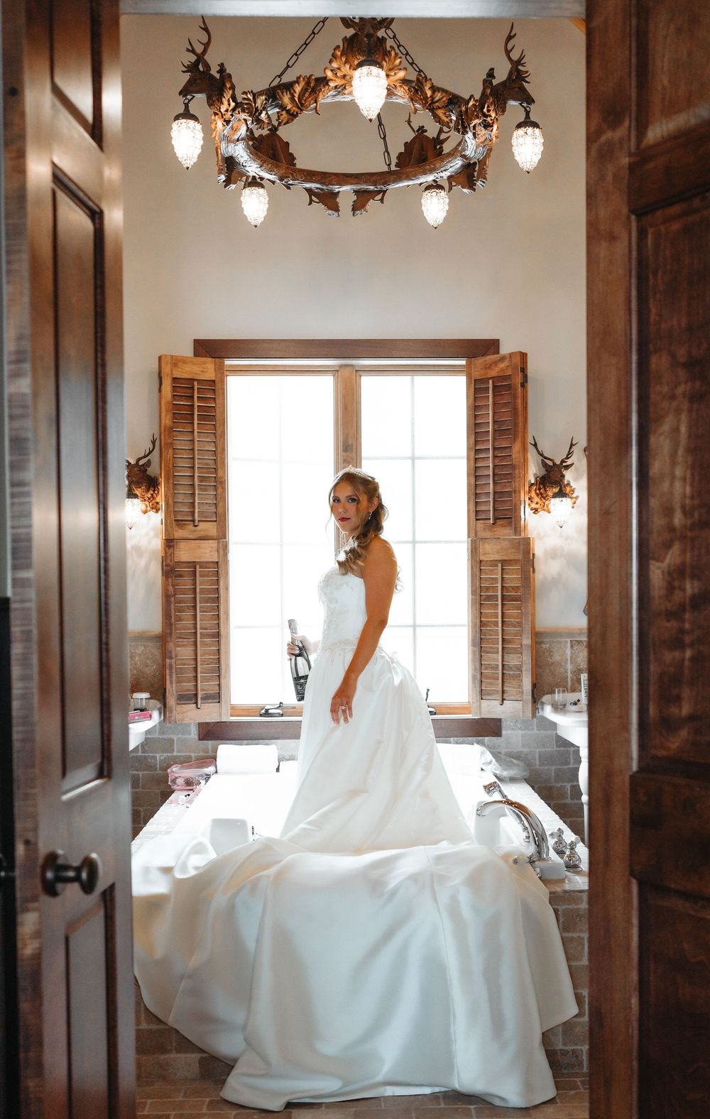 Elegant bride in white gown standing in rustic bathroom with antler chandelier and wooden shutters.