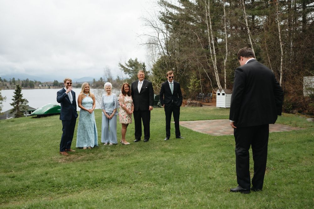 Wedding party gathers on lakeside lawn as groom awaits bride during outdoor ceremony.