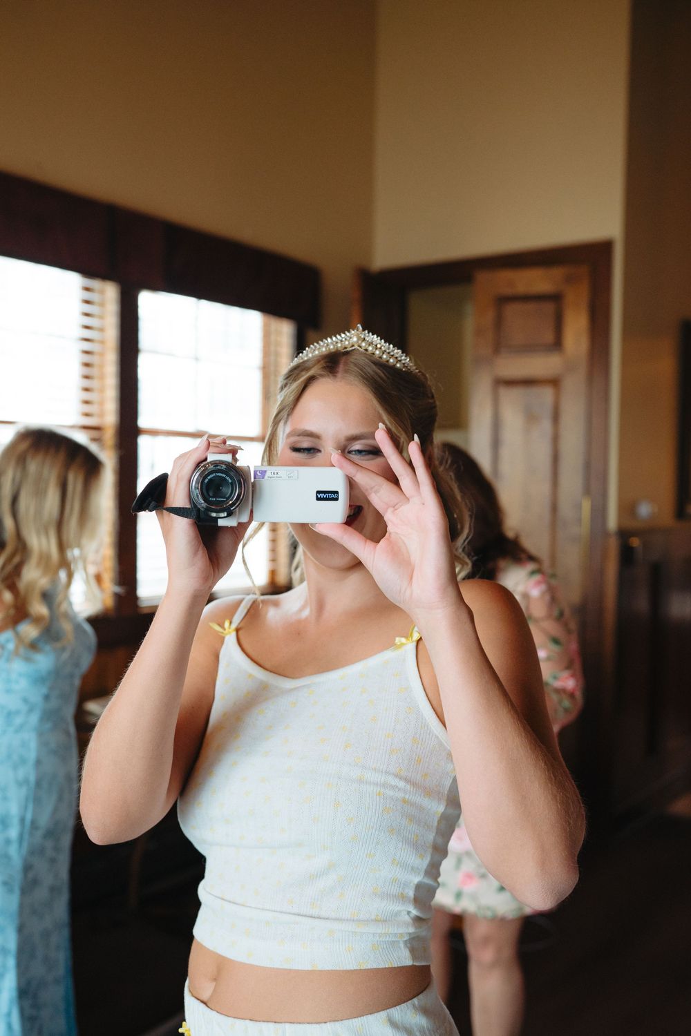 Young woman wearing a tiara films with a handheld video camera indoors at a wedding preparation event.