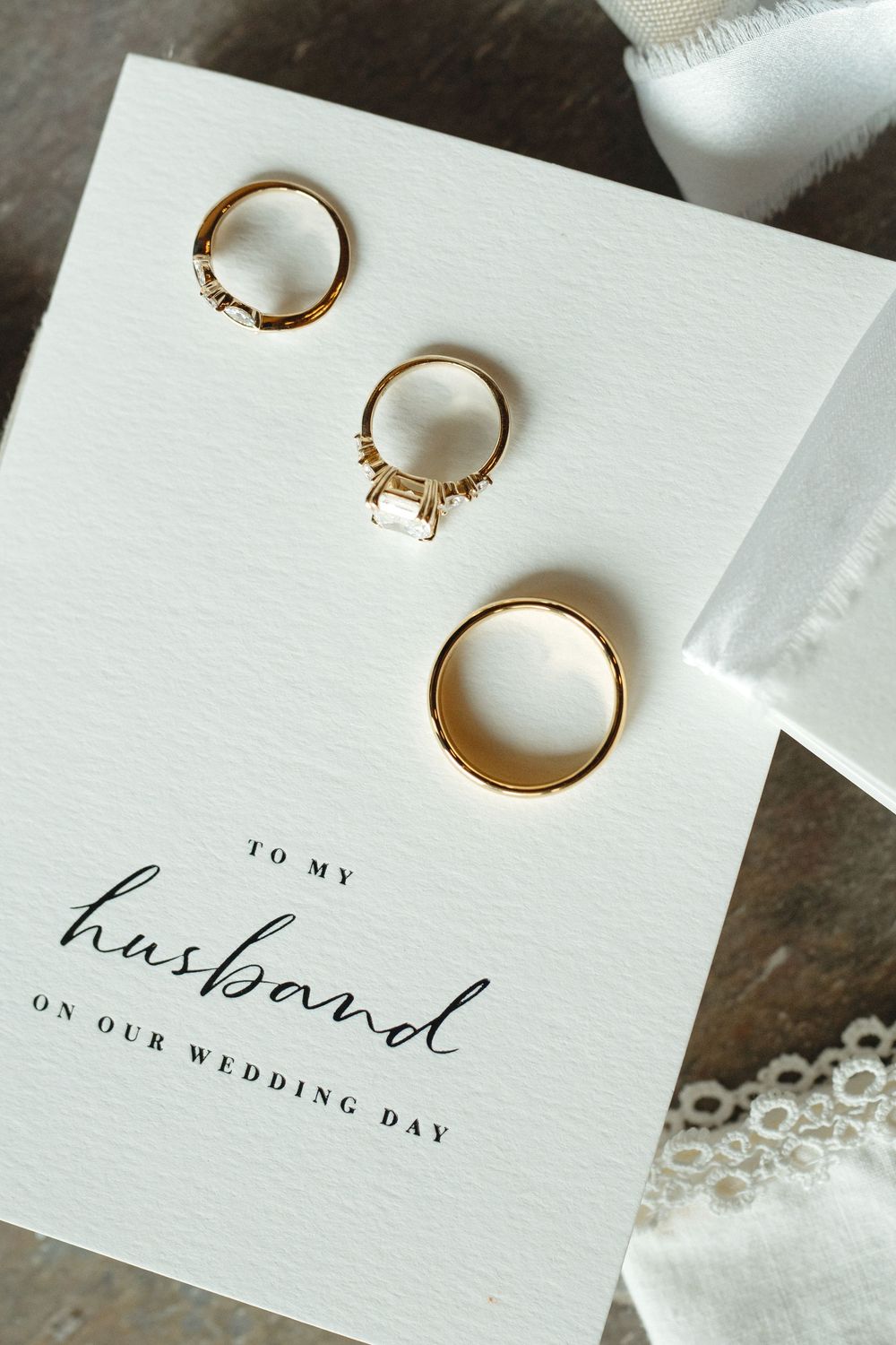 Three gold wedding rings displayed on a white card reading To My Husband On Our Wedding Day with lace and ribbon accents.