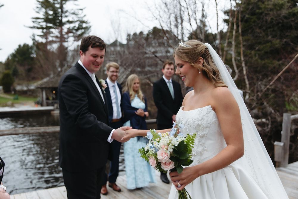 Smiling bride holding floral bouquet exchanges vows with groom on rustic dock near lake.