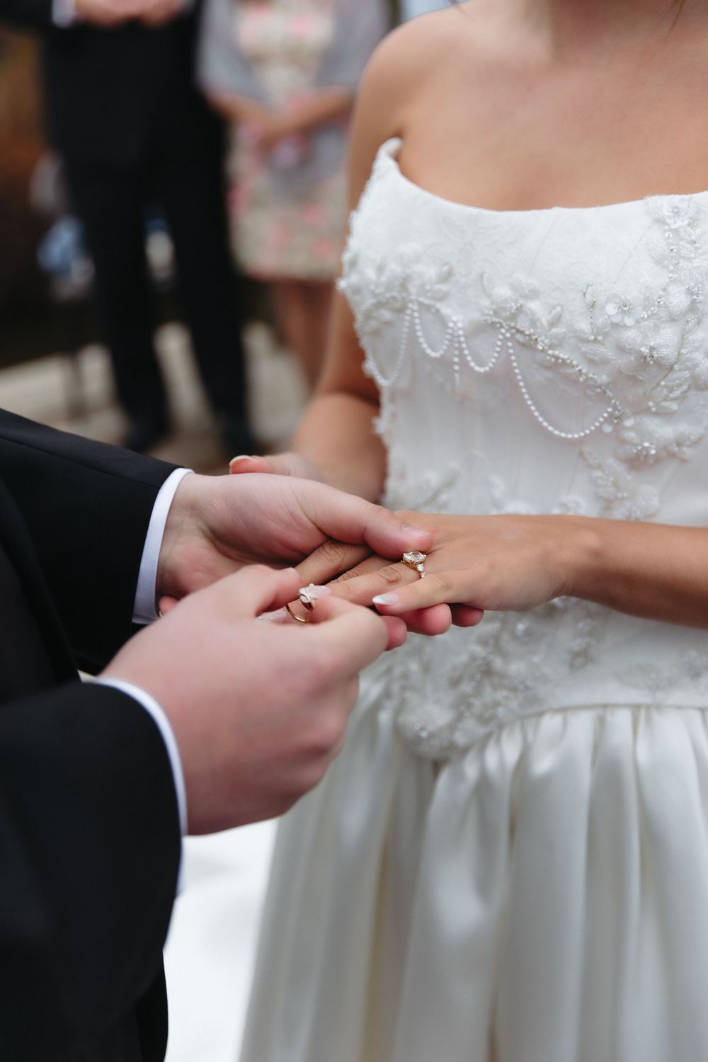 Groom placing wedding ring on brides finger during ceremony, bride wearing elegant white gown.
