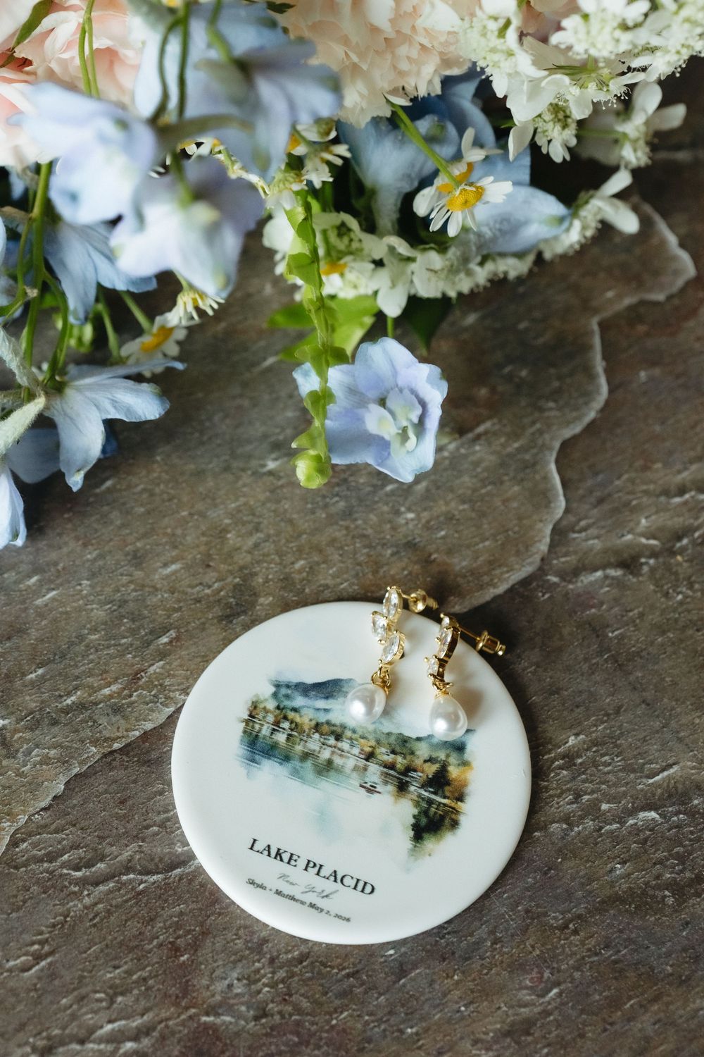 Pearl and gold earrings on a Lake Placid ceramic dish beside a soft blue and white floral bouquet on stone.