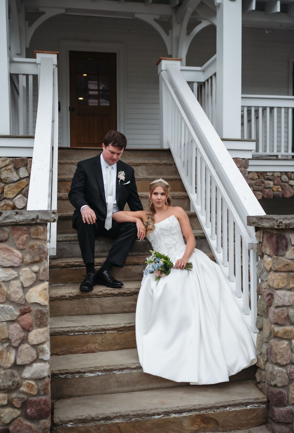 Bride in white strapless gown and groom in black tuxedo sitting on stone steps of a white Victorian porch.