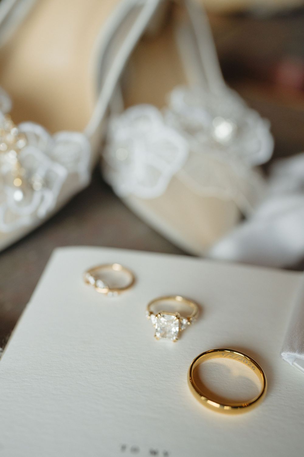 Gold wedding rings and diamond engagement ring displayed on white stationery beside elegant white bridal heels with floral details.
