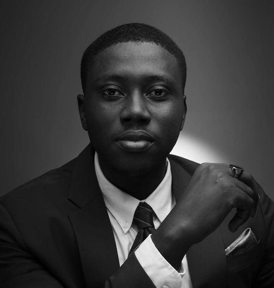 Black and white portrait of a young man in a suit and tie, posing confidently with hand raised to chin.